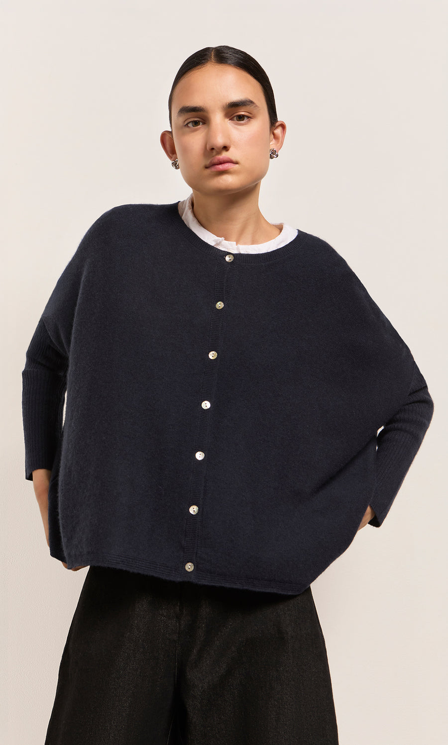 Plümo  Model in Alma Cardigan - Dark Navy over a white shirt, standing against a plain, light-colored background.