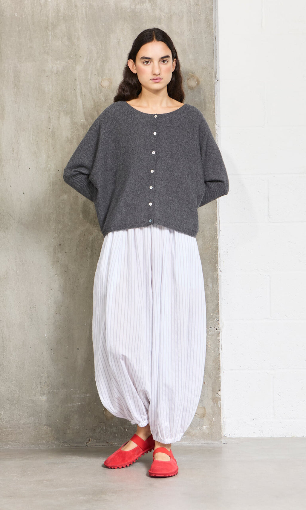 Plümo- A woman in the grey Alma Cardigan, white loose pants, and red shoes stands by a concrete wall.