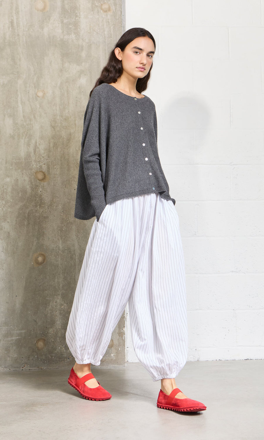 Plümo- Woman in a loose gray sweater and Vento Pants with red shoes stands by a concrete wall.