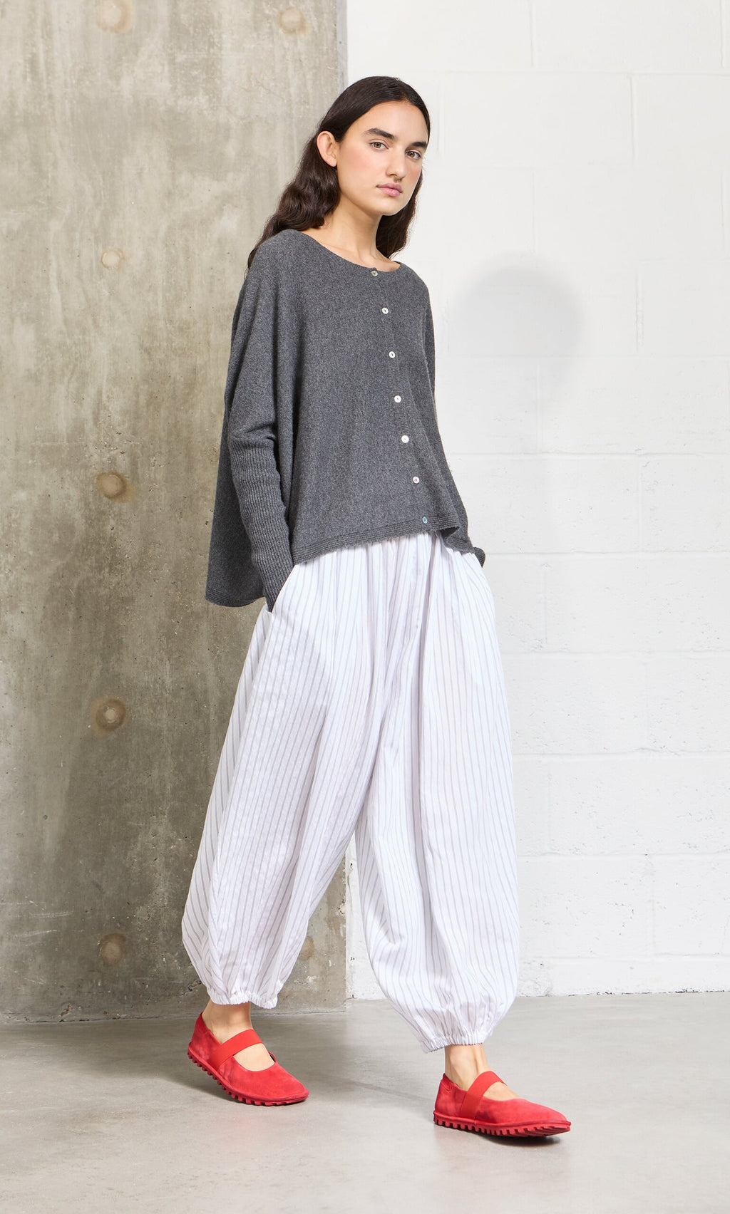 Plümo- Woman in a gray sweater, white striped pants, and Sola Red Flats stands by a concrete and white wall.