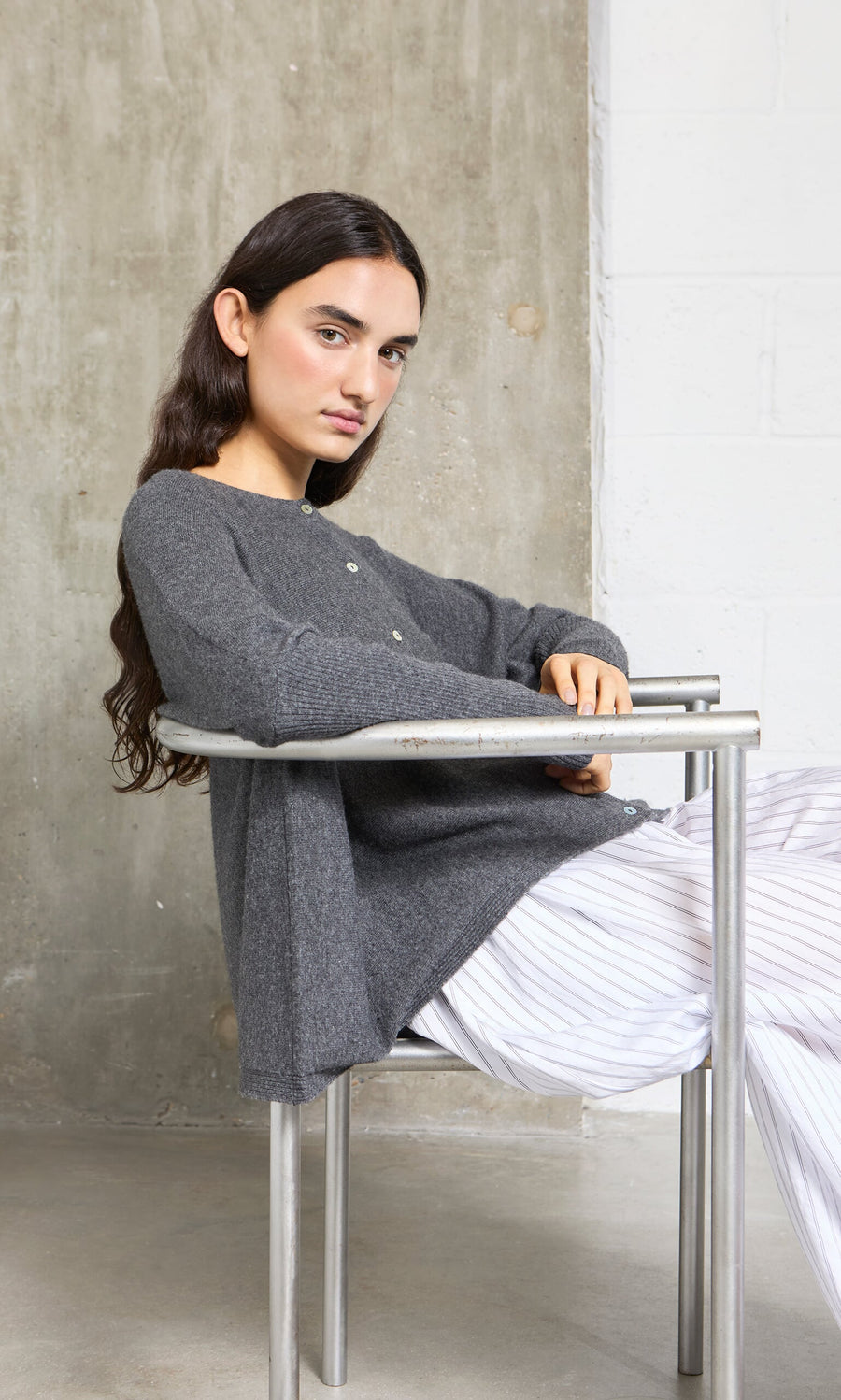 Plümo- Woman with long dark hair sits sideways on a metal chair, wearing the Alma Cardigan - Grey and white striped pants.
