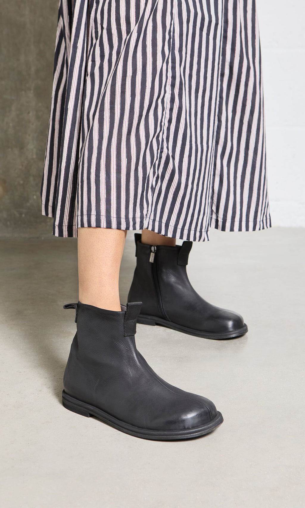Plümo- Person wears Ancona Boots in black waxy leather and a striped skirt, inspired by sleek Danish design, on a light floor.