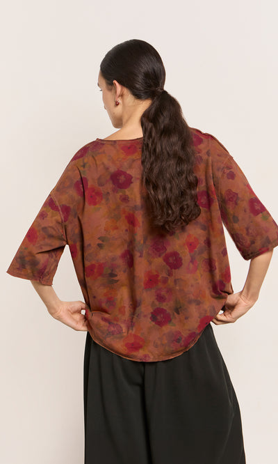 Plümo  Woman with long dark hair in a ponytail wears the Angie Tee, an impressionistic floral brown top, and black pants, back view.