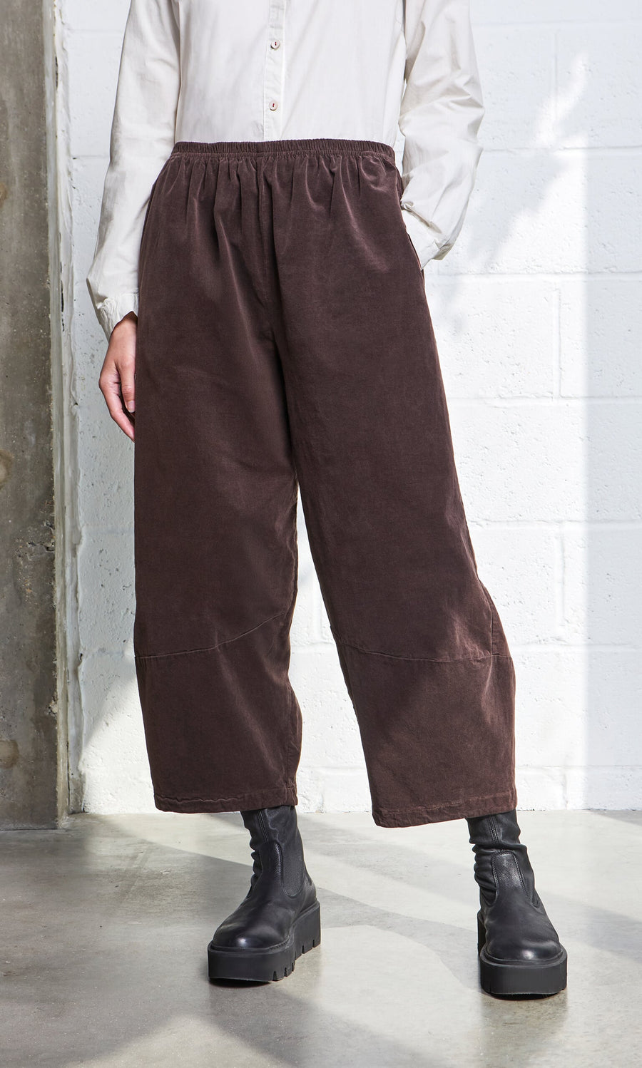 Plümo - Person wearing Anise Pants, black platform boots, and a white button-up shirt standing by a white wall.