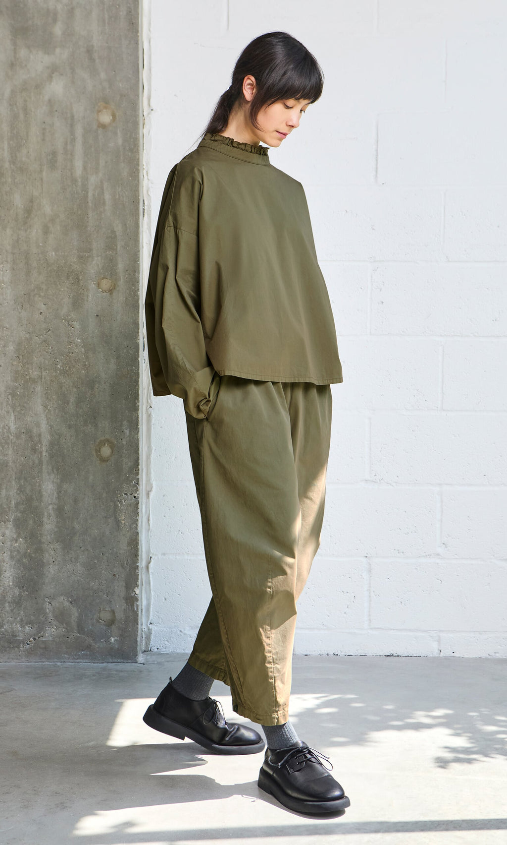 Plümo- Person in olive green Findlay Pants and black shoes stands indoors by a white brick wall and concrete pillar.