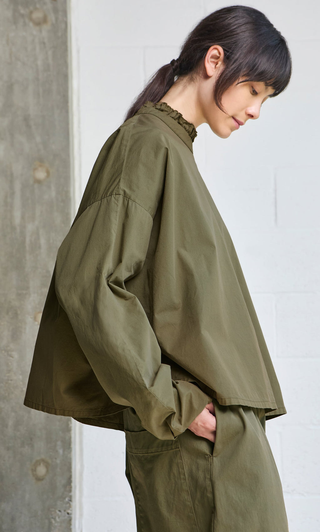 Plümo- Someone in an Olive Ruffle Blouse stands sideways against a light wall, looking down.