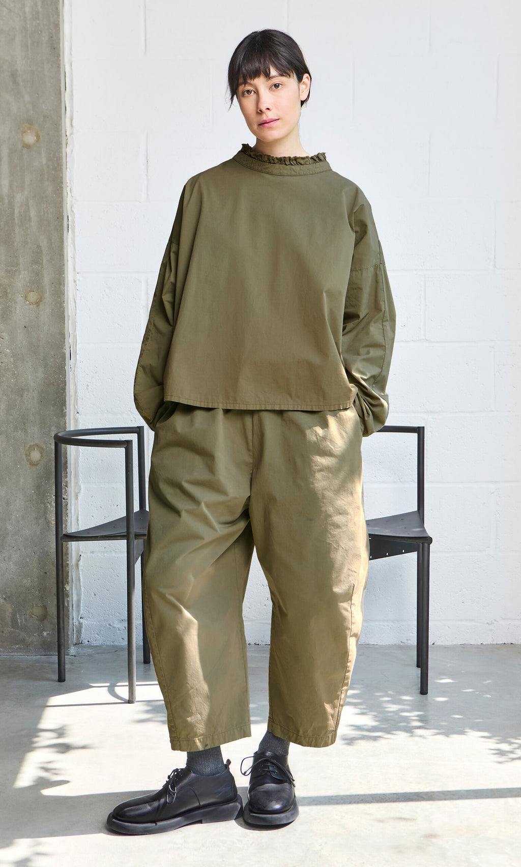 Plümo- Person in an Olive Ruffle Blouse and loose pants, black shoes, standing indoors by a chair with hands in pockets.
