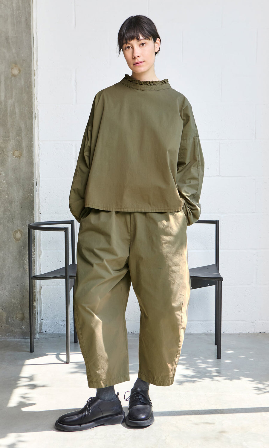 Plümo- Person in an Olive Ruffle Blouse and loose pants, black shoes, standing indoors by a chair with hands in pockets.