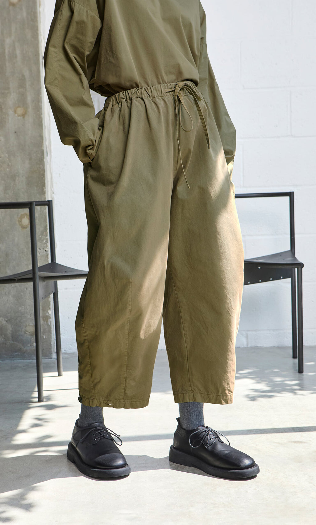 Plümo- Person wears loose olive green Findlay Pants and matching top, black shoes, and gray socks near black chairs indoors.