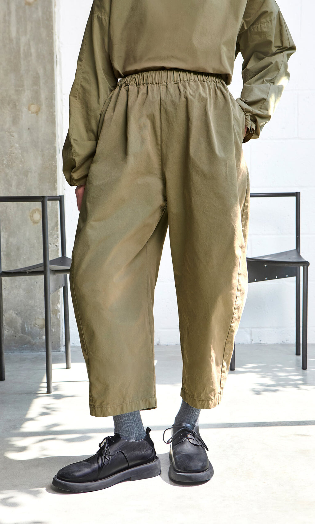 Plümo- Person in olive green Findlay Pants, matching top, gray socks, black shoes stands near black chairs.
