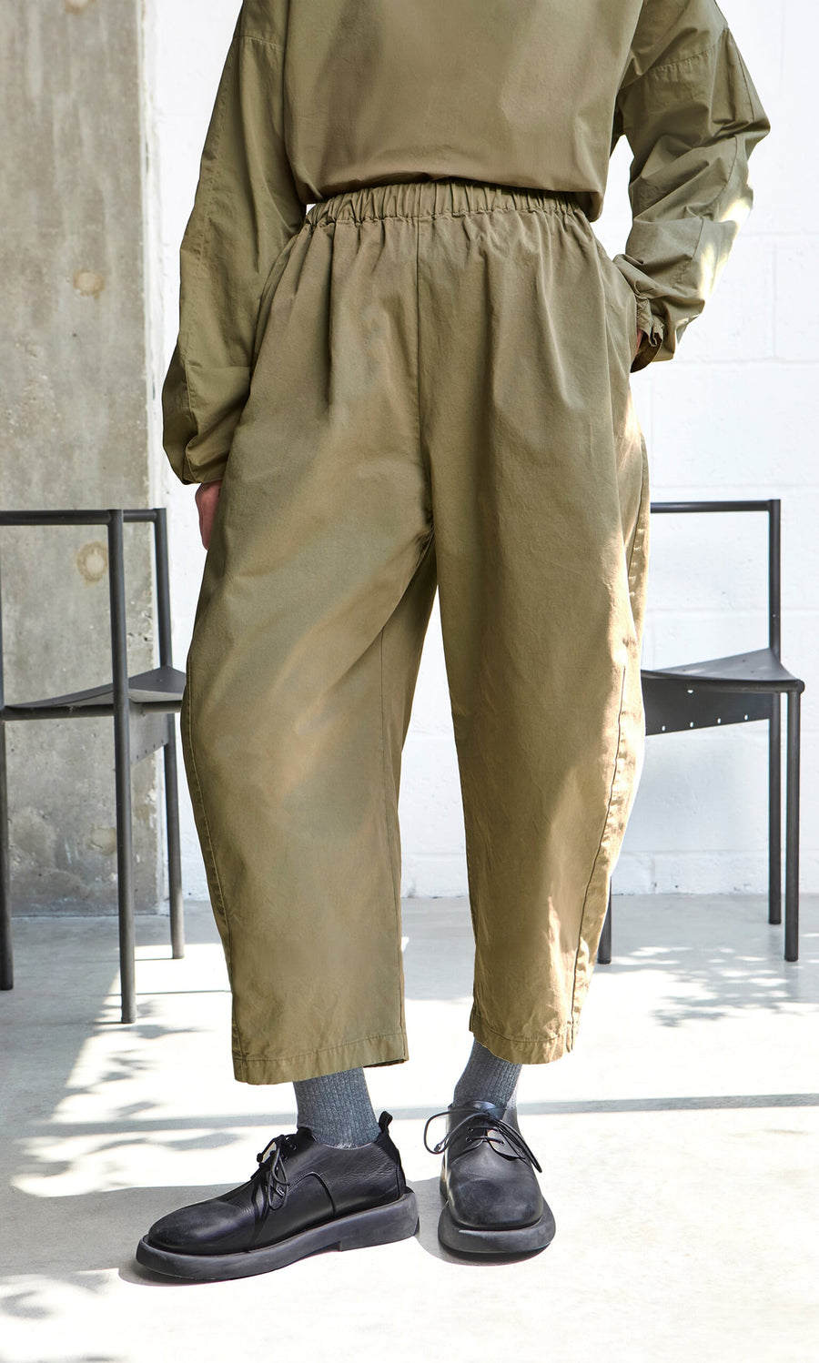 Plümo- Person in olive green Findlay Pants, matching top, gray socks, black shoes stands near black chairs.