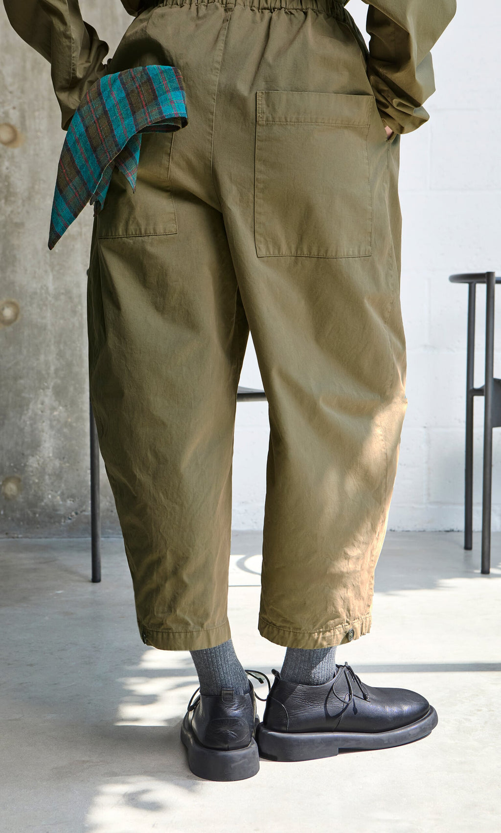 Plümo- Indoors in handcrafted Findlay Pants, gray socks, black shoes, and a plaid cloth tucked in the waistband.