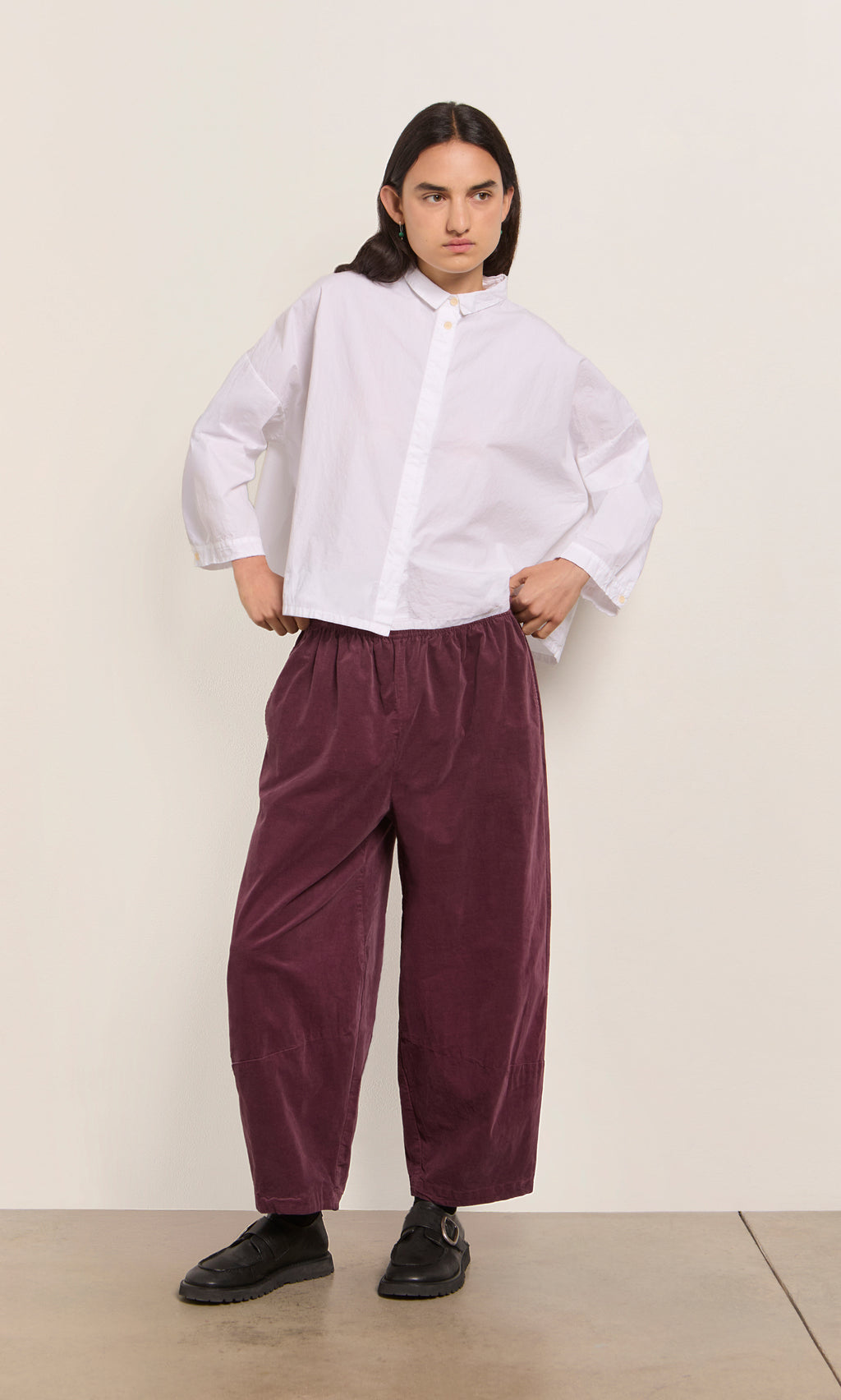 Plümo  Person in a loose white shirt, black shoes, and Architect Pants - Lupins stands against a plain wall.