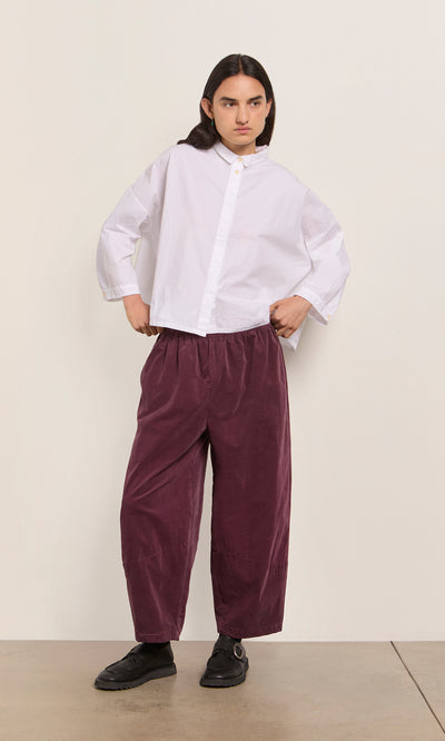 Plümo  Person in a loose white shirt, black shoes, and Architect Pants - Lupins stands against a plain wall.
