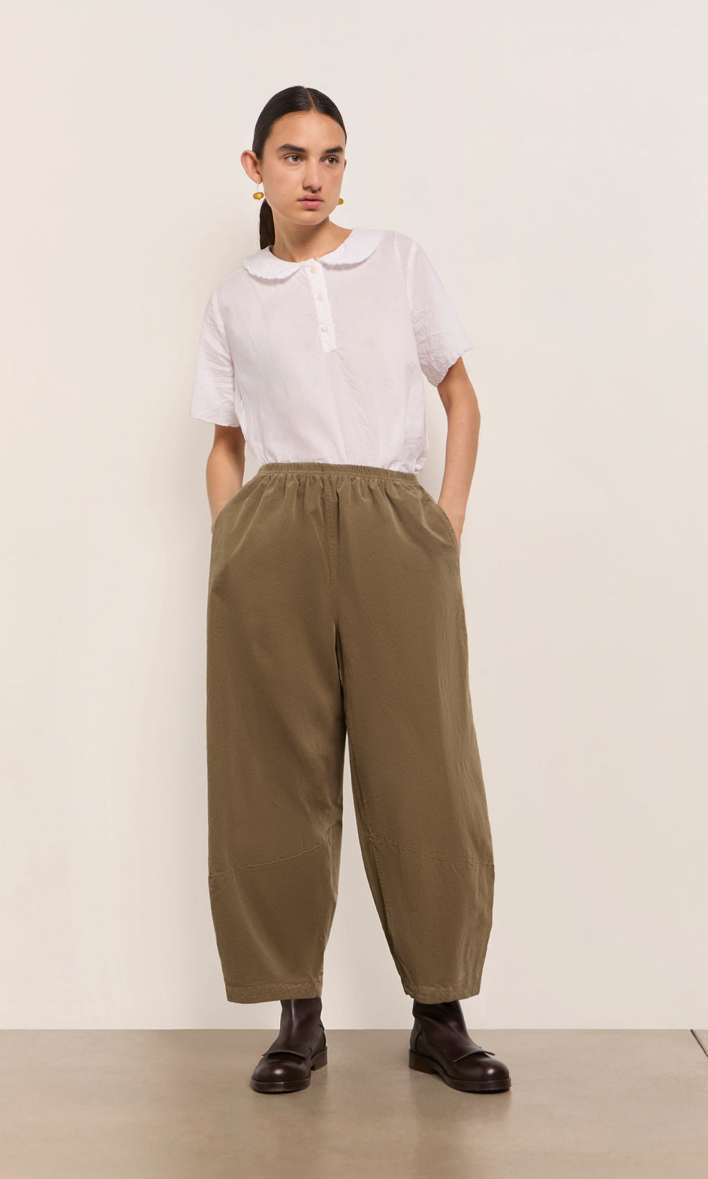 Plümo  Person wears a white collared shirt and Architect Pants- Saddle Brown with dark boots, posing against a plain background.