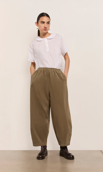 Plümo  Person wears a white collared shirt and Architect Pants- Saddle Brown with dark boots, posing against a plain background.