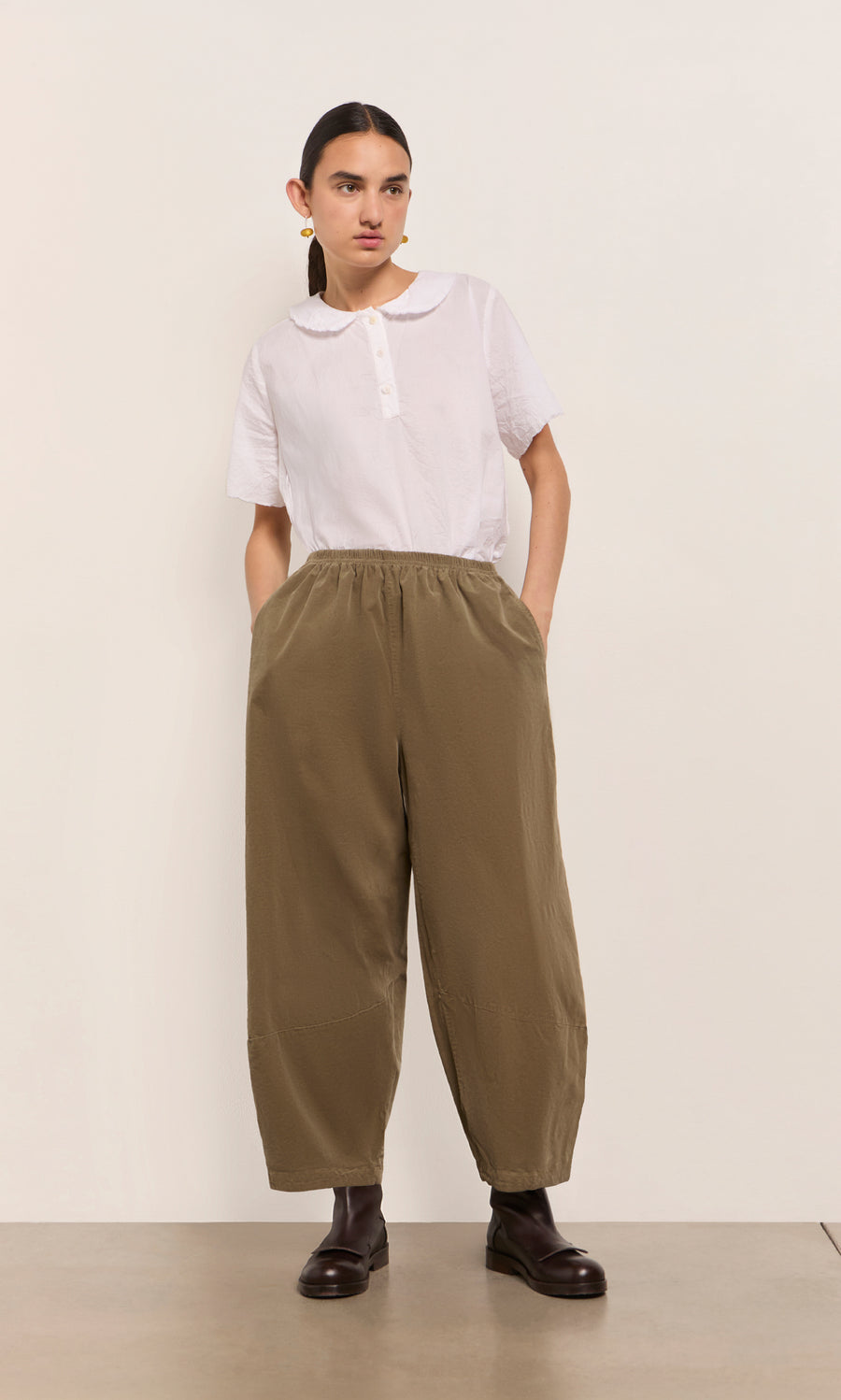 Plümo  Person wears a white collared shirt and Architect Pants- Saddle Brown with dark boots, posing against a plain background.