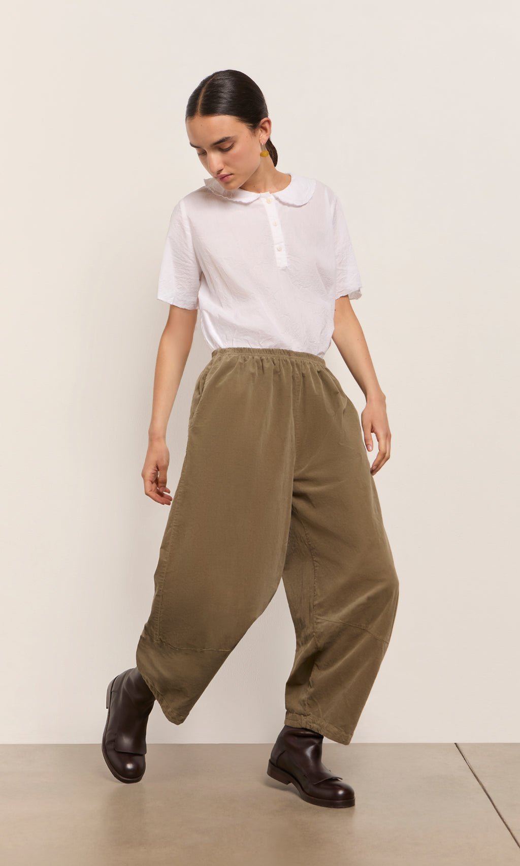 Plümo  A person in a white blouse and Architect Pants—Saddle Brown—plus dark boots stands against a light wall.
