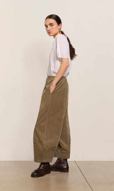 Plümo  Person in white tee, Architect Pants- Saddle Brown, and dark boots stands sideways with hands in pockets.