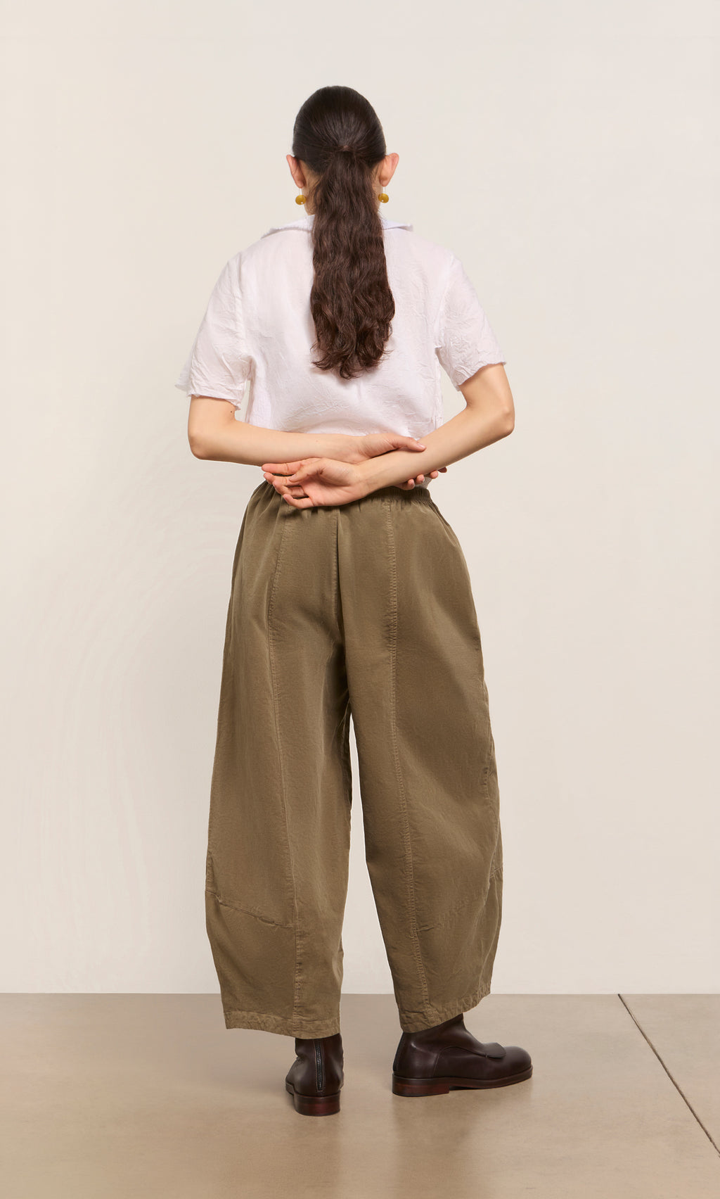 Plümo  Person with long ponytail, white shirt, and Architect Pants in Saddle Brown stands hands behind back, facing away.