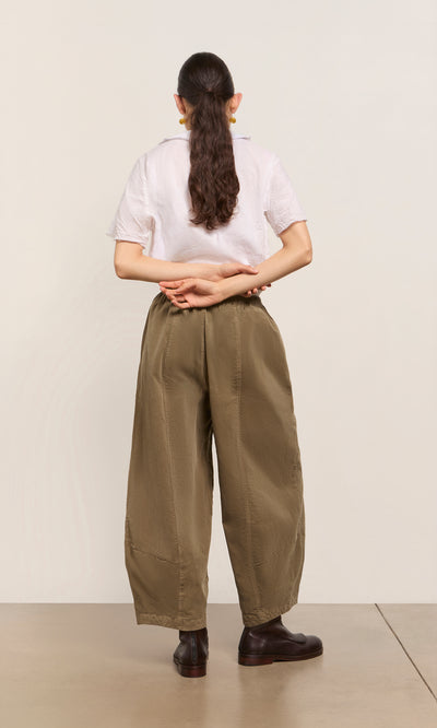 Plümo  Person with long ponytail, white shirt, and Architect Pants in Saddle Brown stands hands behind back, facing away.