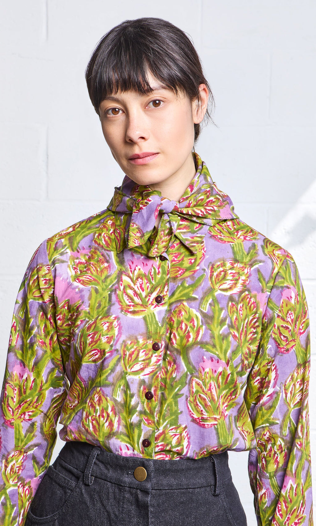 Plümo- A woman wears the Artichoke Shirt—a colorful, block-printed cotton top with a neck bow—paired with dark pants.