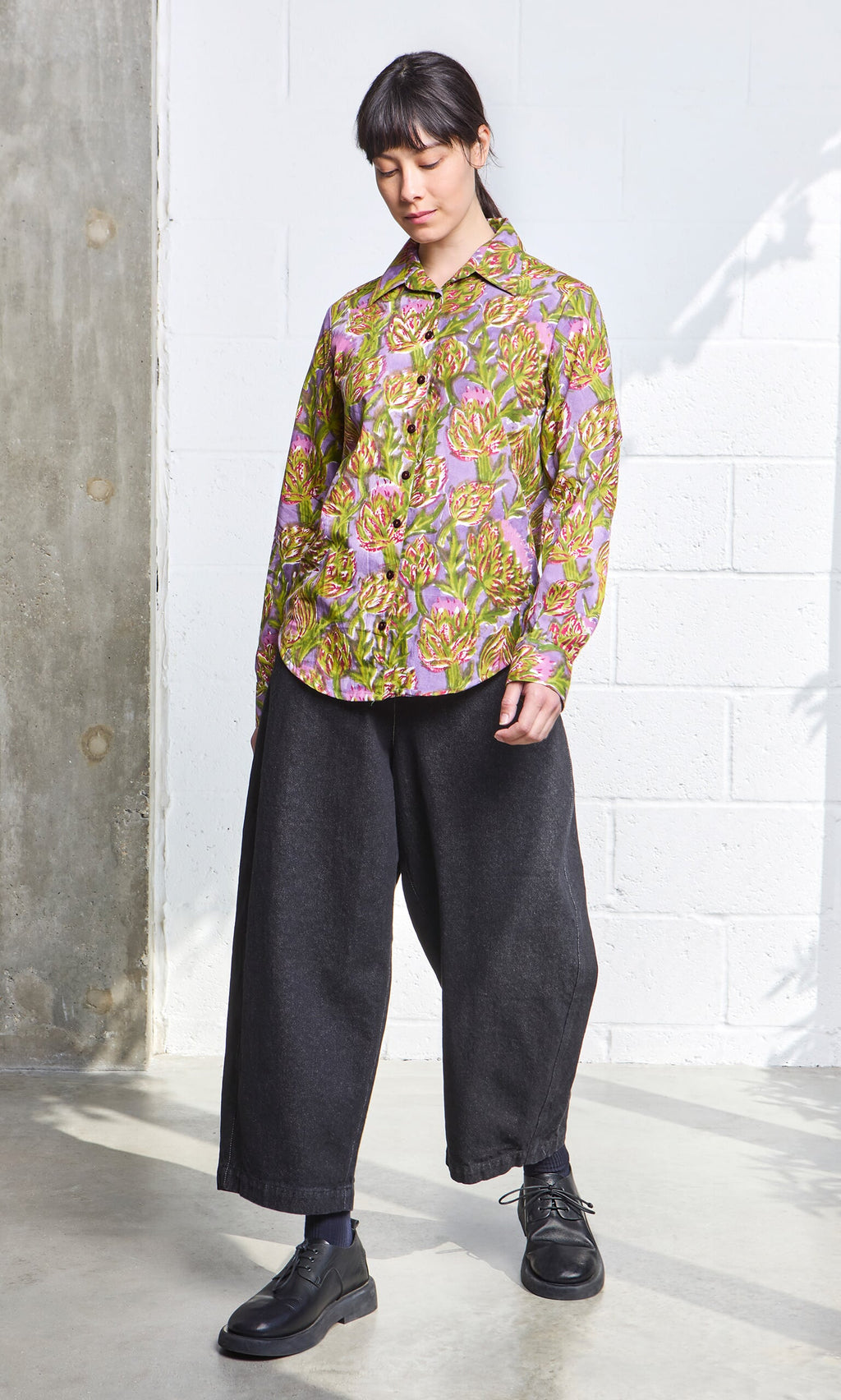 Plümo- Person wears the Artichoke Shirt and wide black pants, standing indoors against a white brick wall.