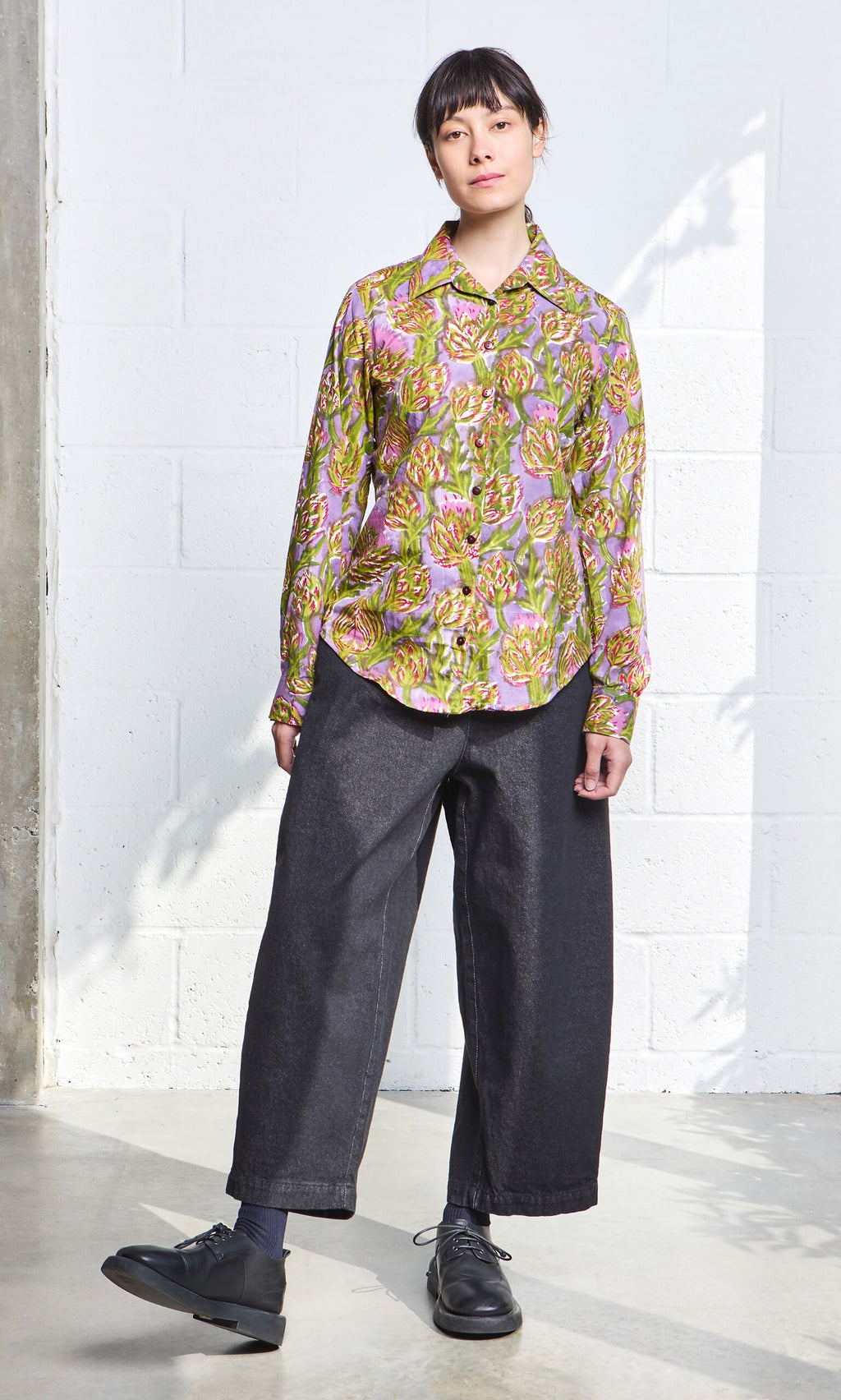 Plümo- Person wearing the Artichoke Shirt stands indoors against a white wall, looking at the camera.