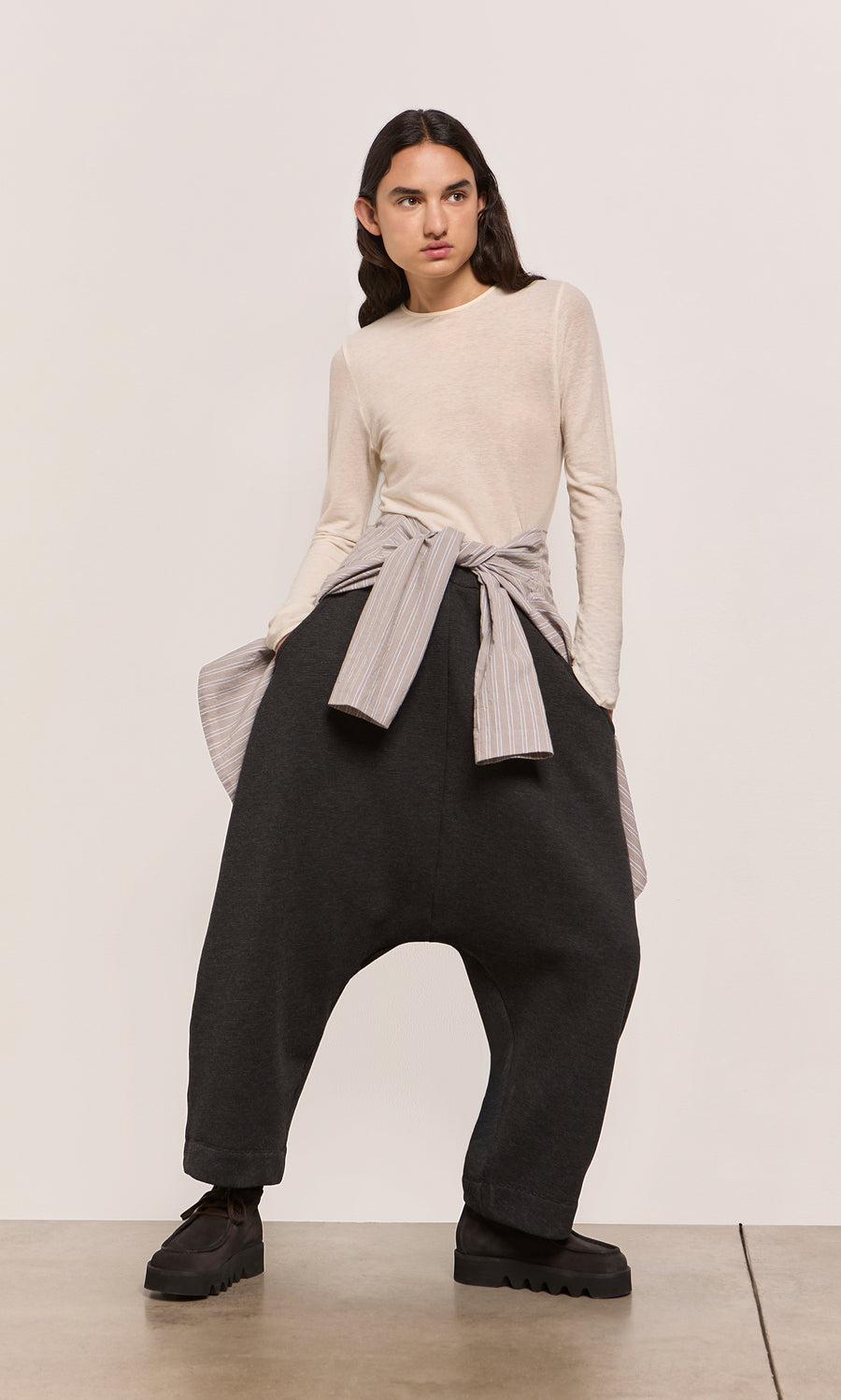 Plümo  Person in Asker Grey Pants, cream long-sleeve shirt tied at waist, standing on a concrete floor.