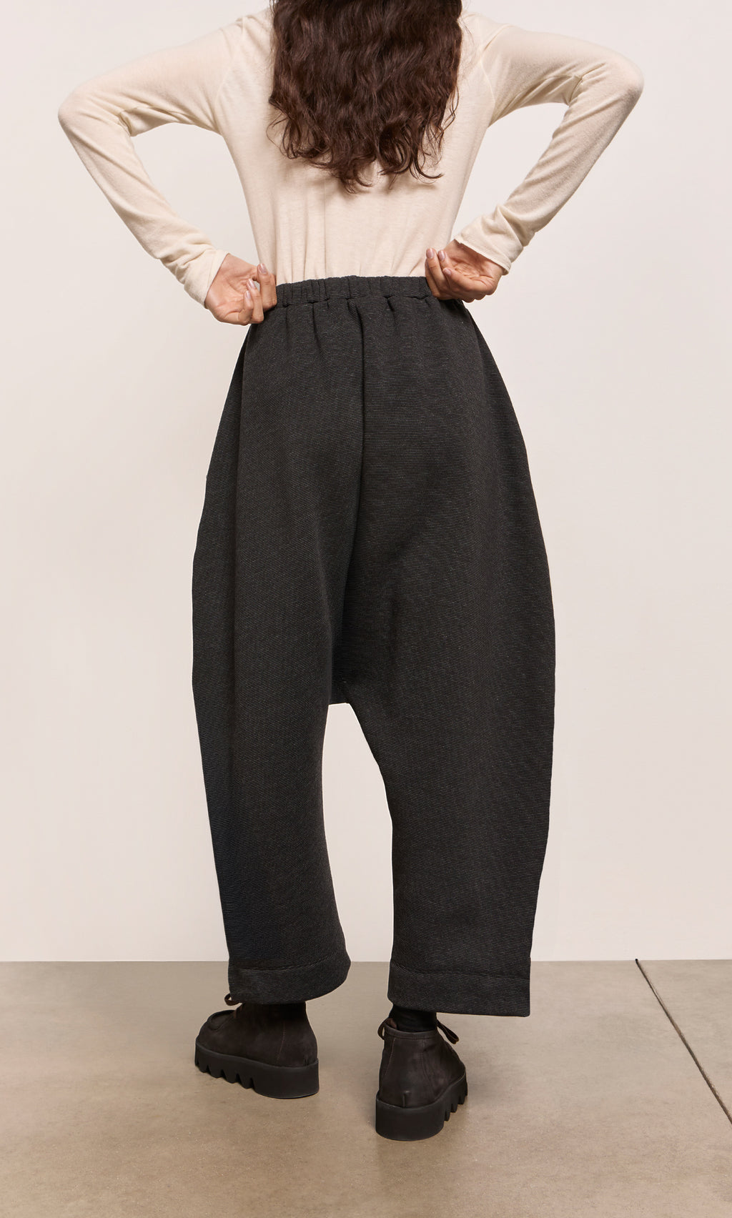 Plümo  Person in beige top and black dropped-crotch silhouette pants stands with hands on hips, facing away, on a concrete floor.