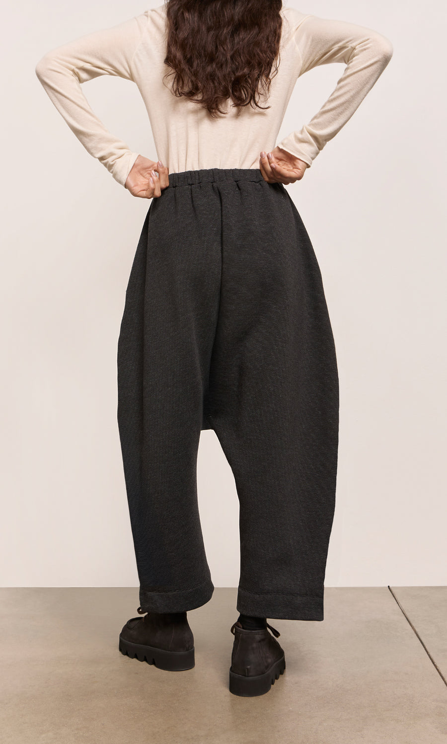 Plümo  Person in beige top and black dropped-crotch silhouette pants stands with hands on hips, facing away, on a concrete floor.