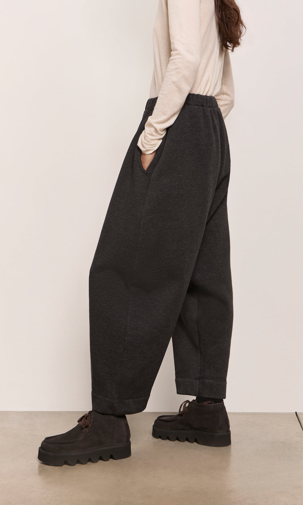 Plümo  Person in Asker Grey Pants, black shoes, and cream top, standing sideways indoors.