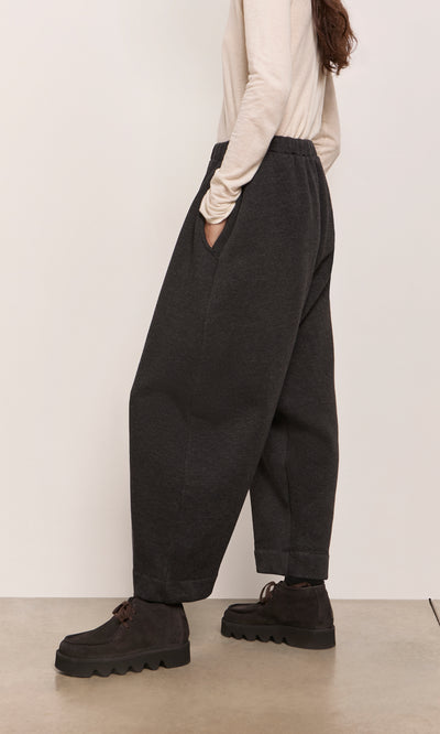 Plümo  Person in Asker Grey Pants, black shoes, and cream top, standing sideways indoors.