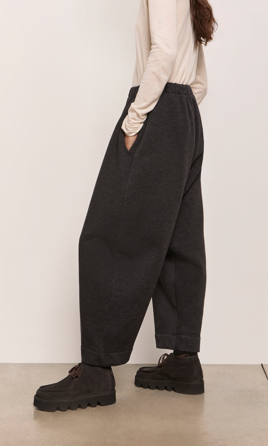 Plümo  Person in Asker Grey Pants, black shoes, and cream top, standing sideways indoors.
