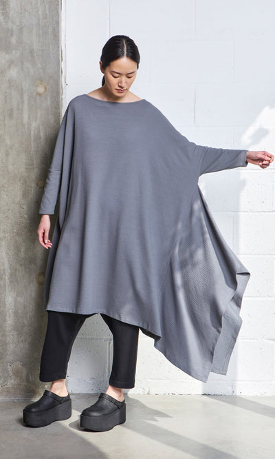 Plümo- Woman in a loose gray tunic, black pants, and Kim Clogs poses by a concrete wall.