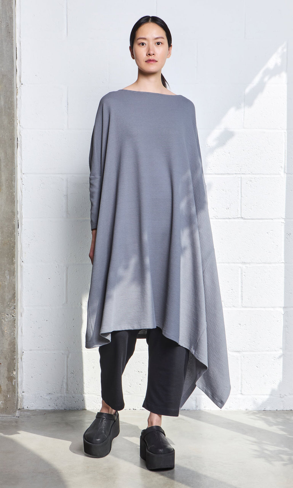 Plümo- A woman in the Asymmetric Dress stands by a white brick wall, paired with black pants and platform shoes.