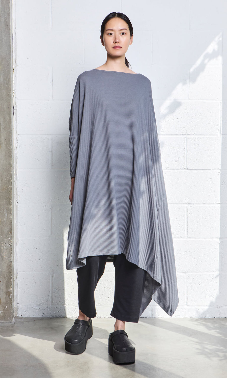 Plümo- A woman in the Asymmetric Dress stands by a white brick wall, paired with black pants and platform shoes.