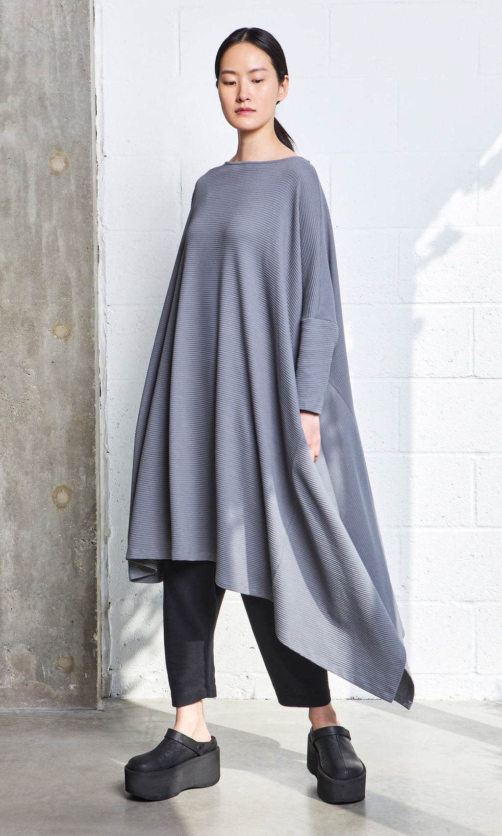 Plümo- Woman wearing the Asymmetric Dress in gray organic cotton over black pants and platform shoes, standing by a white wall.