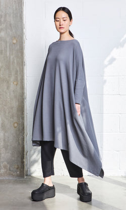 Plümo- Woman wearing the Asymmetric Dress in gray organic cotton over black pants and platform shoes, standing by a white wall.
