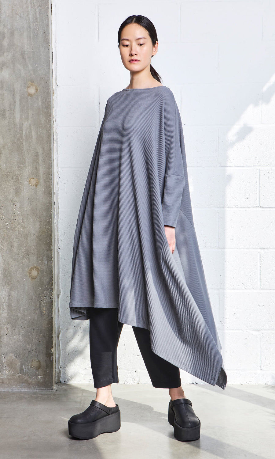 Plümo- Woman wearing the Asymmetric Dress in gray organic cotton over black pants and platform shoes, standing by a white wall.