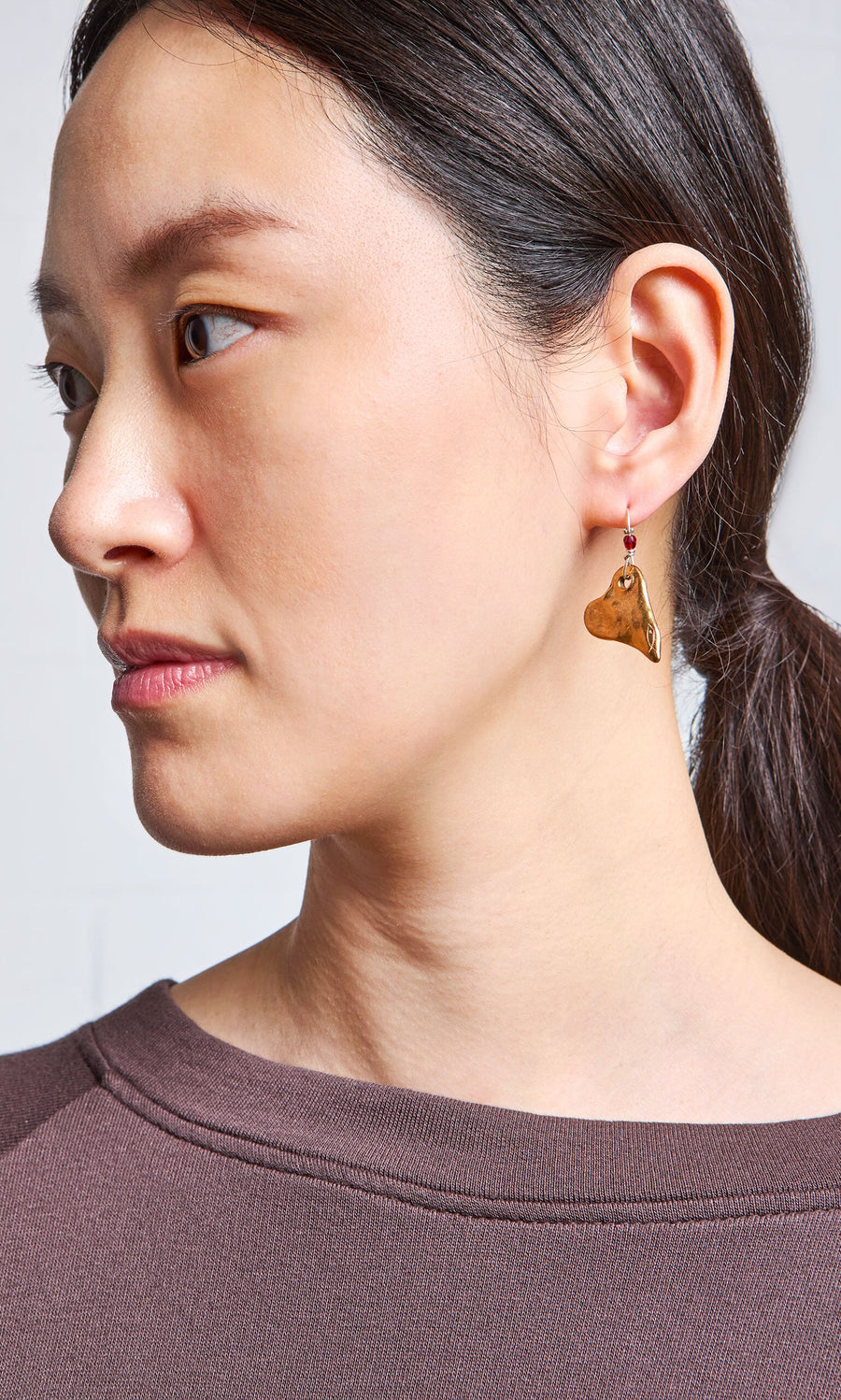 Plümo- Woman with dark hair in a ponytail, wearing a brown shirt and Bacio Earrings, looks to the left.