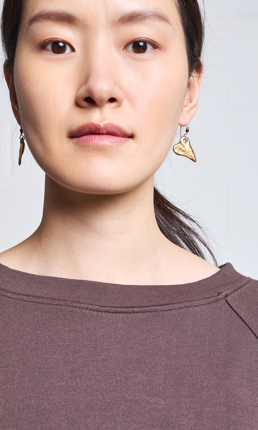 Plümo- Woman with straight dark hair in a ponytail wears Bacio Earrings, handmade gold abstracts, and a brown top.