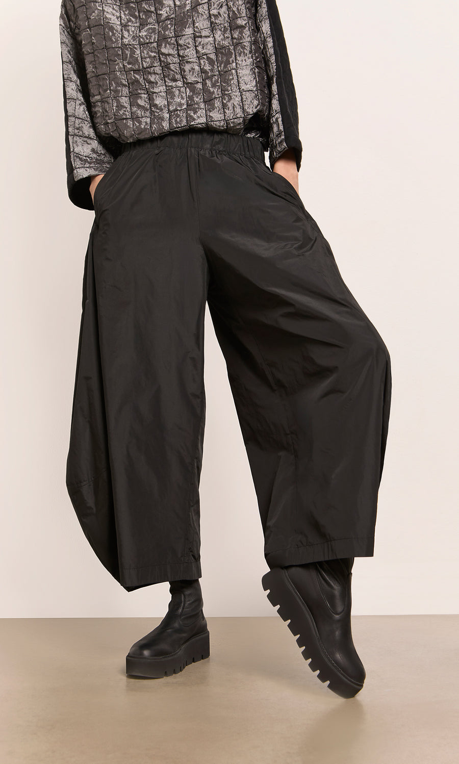 Plümo  Model in Hooley Pants, chunky black boots, and a textured gray top stands with one leg raised.