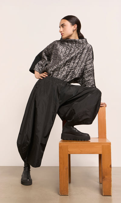 Plümo  Woman in a gray textured top and Hooley Pants poses on a wooden chair, looking to the side.