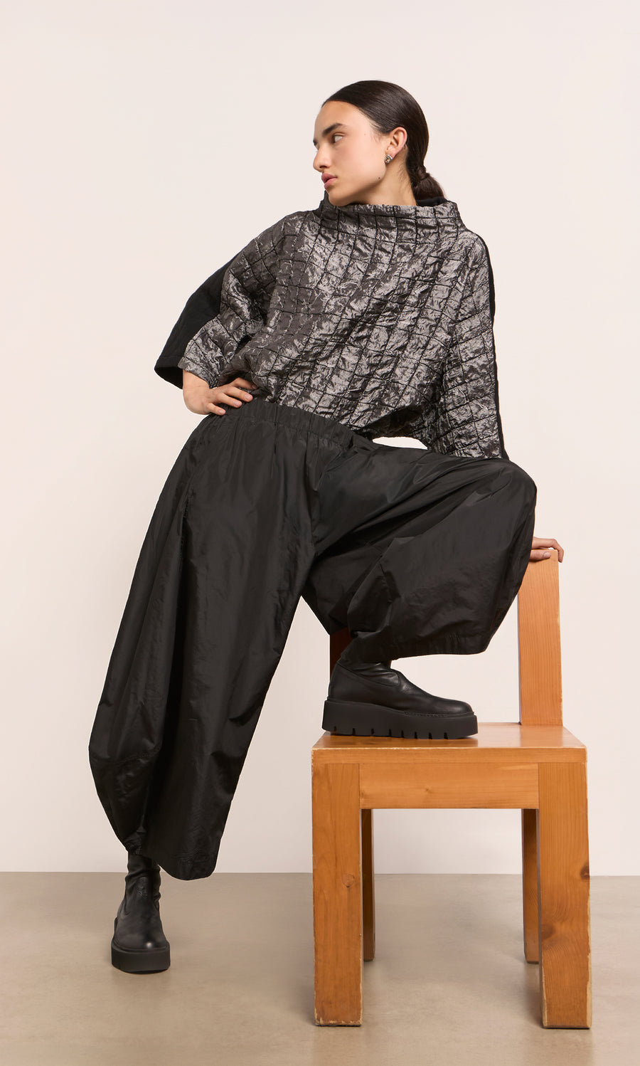 Plümo  Woman in a gray textured top and Hooley Pants poses on a wooden chair, looking to the side.