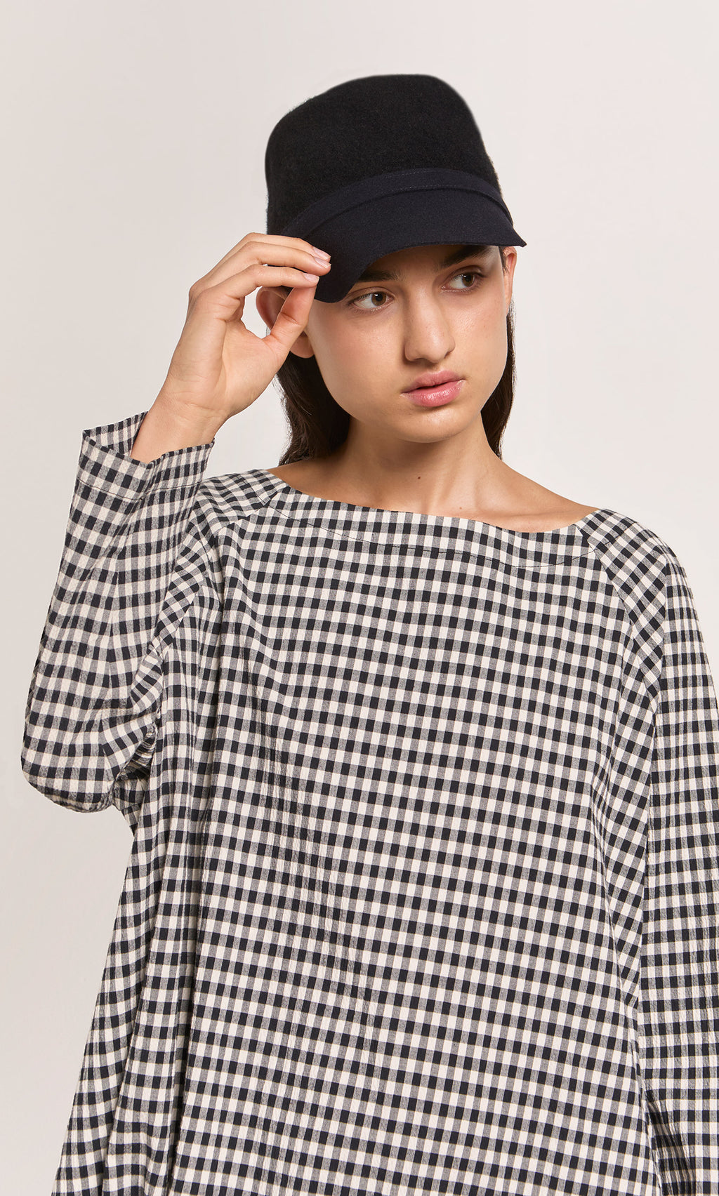 Plümo  Woman in a Baseball Cap and loose black-and-white checkered top looks to the side against a plain background.