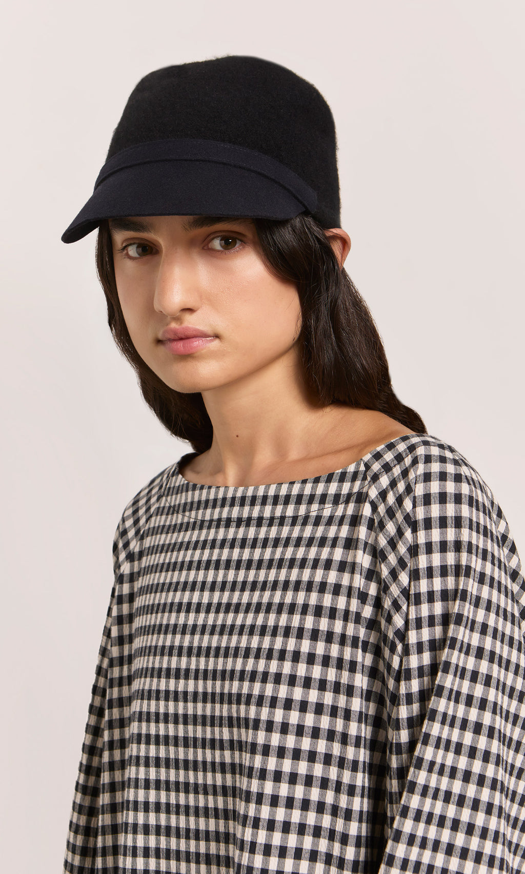 Plümo  Woman in a Baseball Cap and checkered top looks at the camera with a neutral expression.