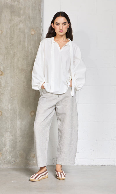 Plümo- Woman in a white blouse, wide-leg pants, and Basque Espadrilles stands by a concrete and white wall.