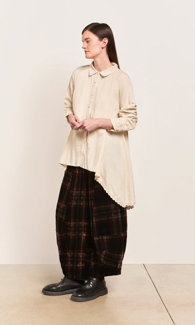 Plümo  A woman in a cream shirt and Carson Tartan Pants stands indoors on a light floor, facing left.