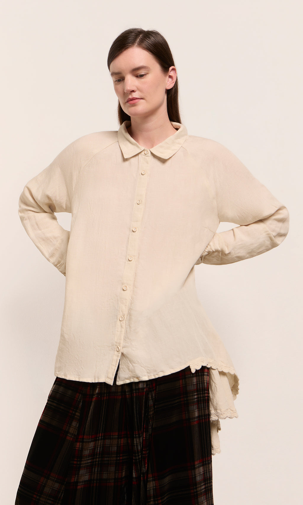 Plümo  Woman wears Beeson Shirt - Oat with a black and red plaid skirt, standing against a plain background.